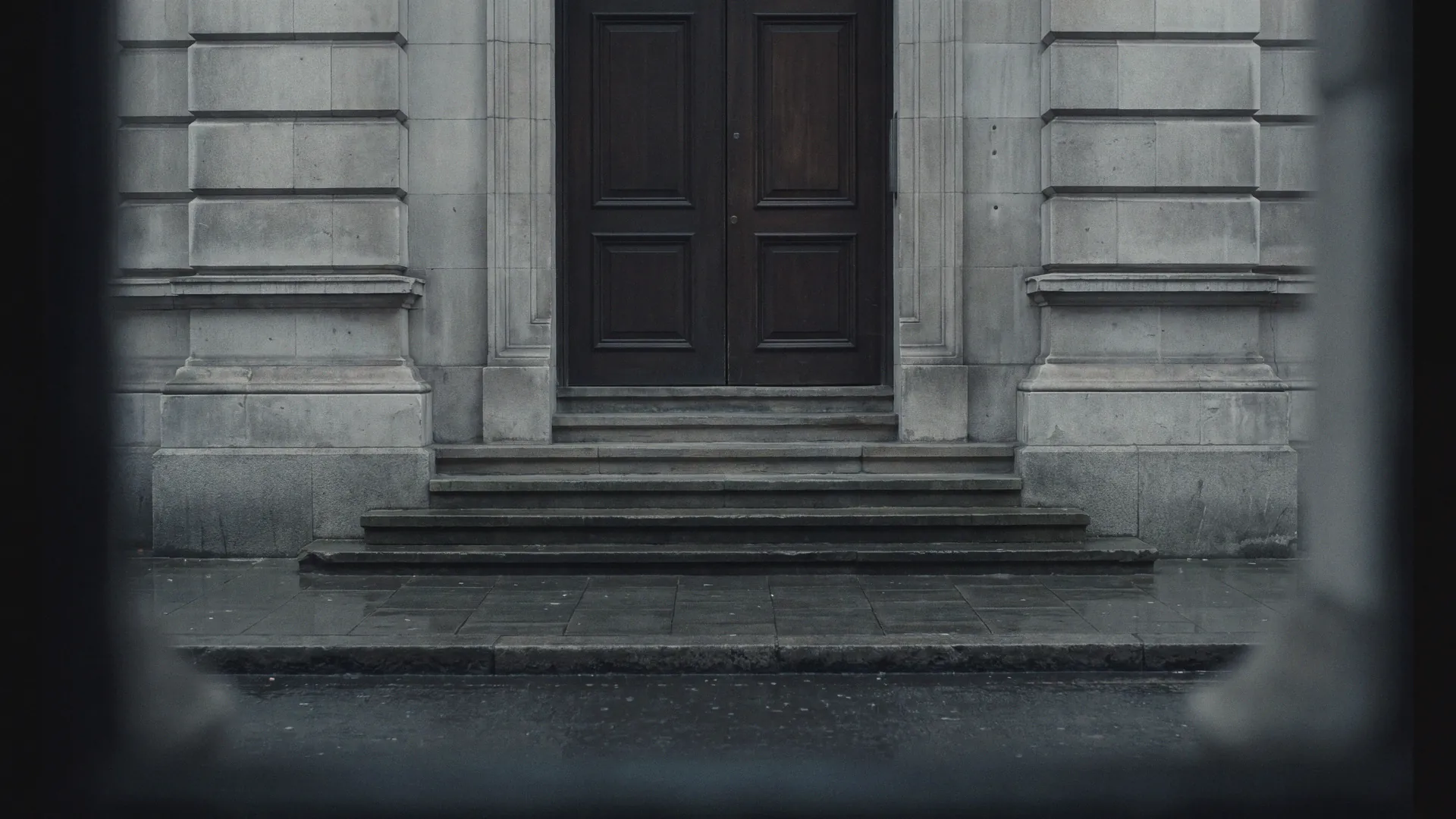 The stone entrance of a London office building on an overcast day, dark door and wet pavement, heavily defocused.
