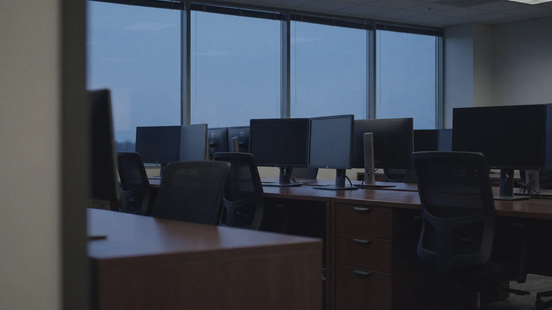 An open-plan office at dusk, empty desks and dark monitors against a city window, heavily defocused.